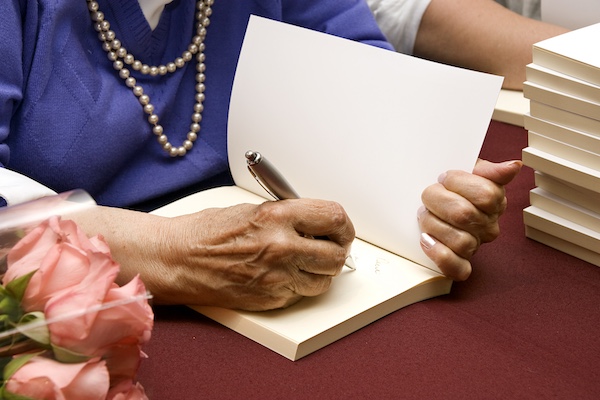 an older woman, wearing a purple sweater and a pearl necklace, signing the inside of a book
