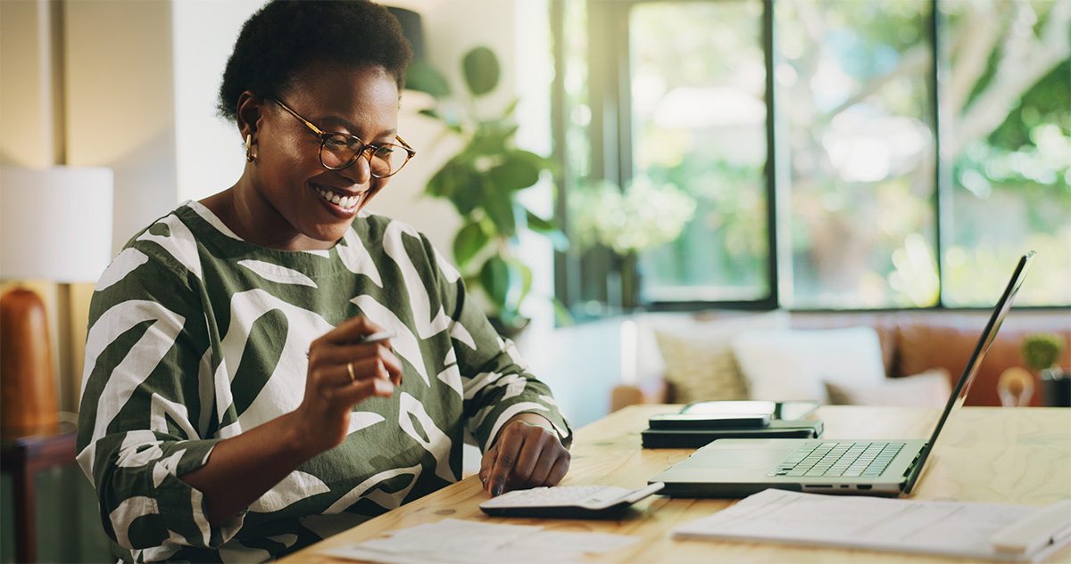 A woman sitting at a wooden table with a laptop, calculator and note pad. She is smiling while she works